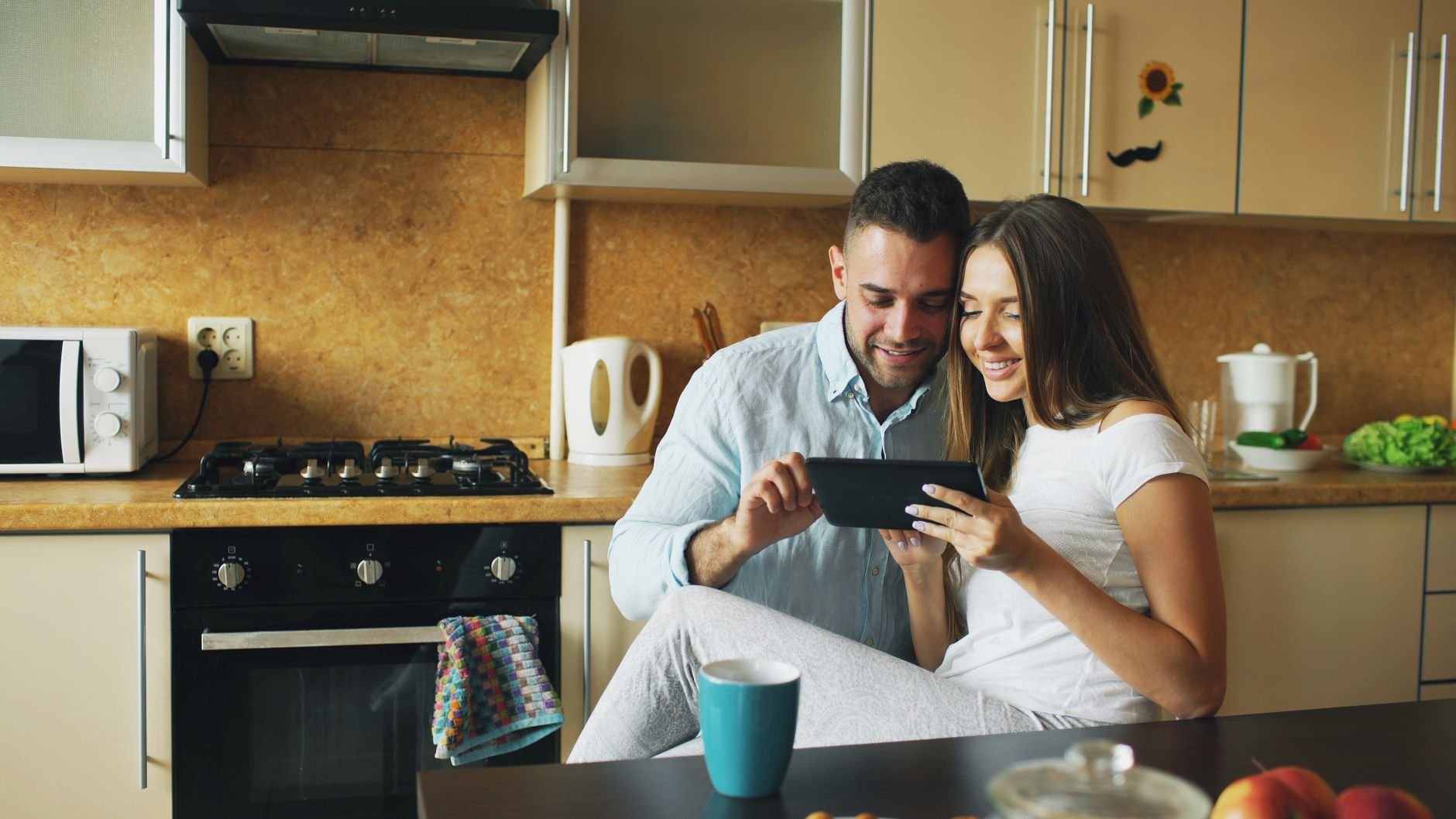 Happy couple using a tablet in a modern kitchen setting, surrounded by cosy and warm decor.